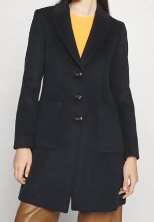 Femme portant un manteau noir à simple boutonnage avec trois boutons, des poches plaquées, par-dessus un haut orange et un pantalon marron.