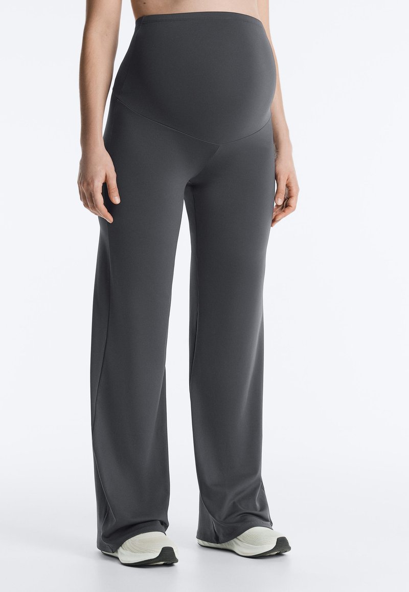 Personne enceinte portant un pantalon de maternité gris foncé avec un large panneau au niveau du ventre et des baskets blanches, debout devant un fond uni.