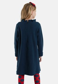 Mädchen mit langen Haaren, das ein marineblaues Kleid mit rüschenartig gestaltetem Kragen und rote Leggings mit großen marineblauen Punkten trägt, mit dem Rücken zur Kamera gewandt.
