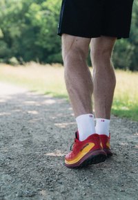 Baskets de course rouges et jaunes avec des accents noirs, portées avec des chaussettes blanches, sur un chemin en gravier. Muscles des jambes visibles et shorts de sport.