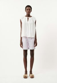 Weiße ärmellose Bluse mit einem Schlüssellochausschnitt, kombiniert mit gestreiften Shorts in mehreren Farben. Das Modell trägt braune Sandalen.