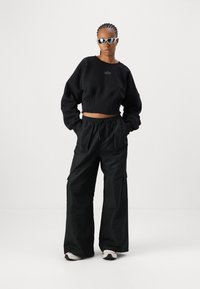 Svart kort sweatshirt med et svakt logo, kombinert med løstsittende svart cargo-bukser som har sidelommer. Hvite og svarte sko.