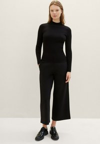 Crna rebrasta turtlenecka majica u kombinaciji s crnim širokim hlačama. Odjevna kombinacija je upotpunjena sjajnim crnim cipelama na vezivanje.