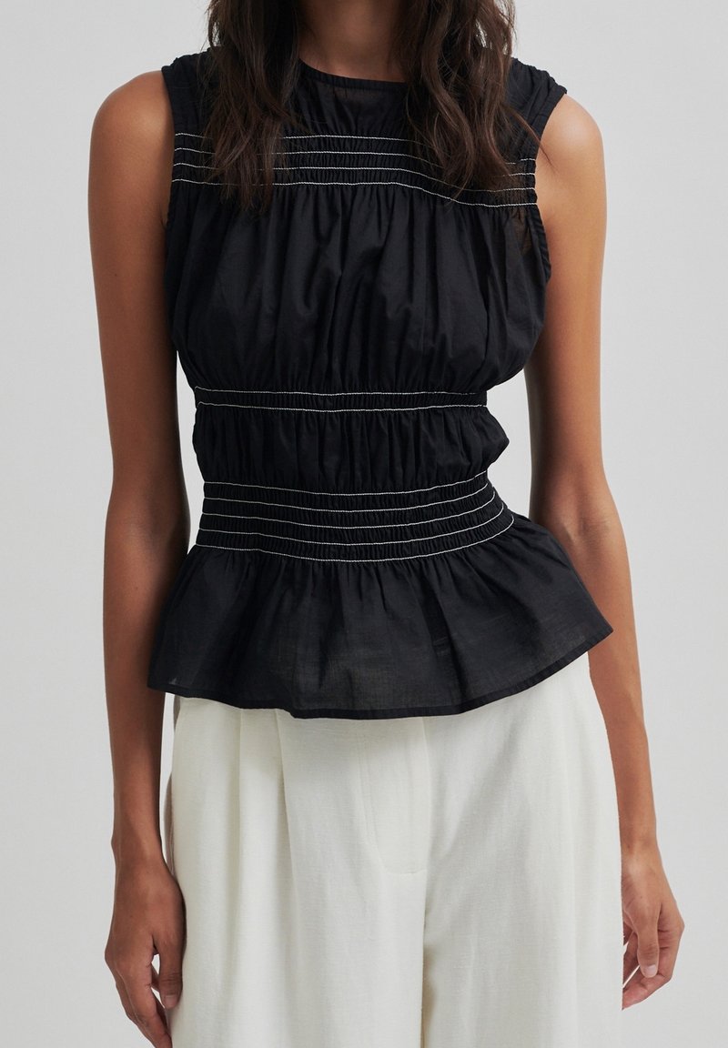 Blouse noire sans manches avec coutures horizontales blanches, taille froncée et ourlet à basque portée avec un pantalon blanc sur une personne.