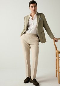 Hellolivfarbener Blazer über einem weißen Hemd, kombiniert mit beigen Hosen und dunkelbraunen Loafers. Einfaches Design, glatte Textur und neutrales Farbschema.