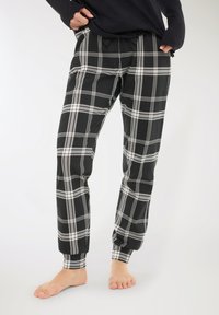 Persona che indossa pantaloni a quadri bianchi e neri con polsini elasticizzati e tasche, in piedi a piedi nudi su uno sfondo neutro.