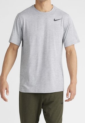 Homme portant un t-shirt Nike à manches courtes gris clair et un pantalon Nike vert foncé, debout avec les bras détendus le long du corps.