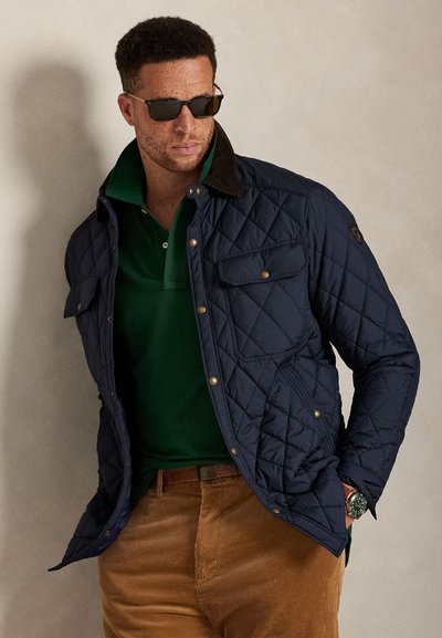 Μπουφάν σε σκούρο μπλε με καπιτονέ ύφασμα, γιακά από κοτλέ, μπροστινές τσέπες και χρυσά κουμπώματα, φορεμένο πάνω από πράσινη πόλο μπλούζα και καφέ παντελόνι από κοτλέ.