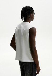 Hvid ærmeløs herreskjorte med en afslappet pasform, blødt bomuldsstof og en sømdetalje på ryggen. Kombineret med mørke jeans.