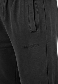 Black joggers made of soft fabric, featuring side pockets and a textured logo detail. The design includes an elastic waistband for comfort.