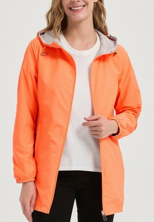 Femme portant une veste à capuche orange vif par-dessus une chemise blanche et un pantalon noir, souriante avec une main près de la fermeture éclair.