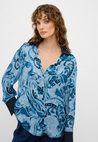 Blusa blu a fantasia con design floreali ondulati, apertura frontale con bottoni e colletto scuro a contrasto. Maniche lunghe con dettagli ai polsini.