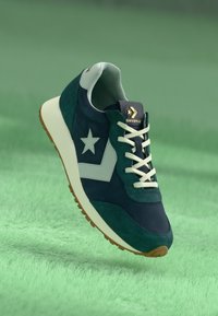 Zaļš un tumši zils zamšādas sporta apavu modelis ar balto zvaigznes logo, krēmbaltiem šņorēm un brūnu gumijas zoli, paredzēts ikdienas valkāšanai.