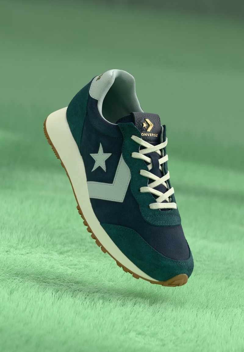 Zaļš un tumši zils zamšādas sporta apavu modelis ar balto zvaigznes logo, krēmbaltiem šņorēm un brūnu gumijas zoli, paredzēts ikdienas valkāšanai.