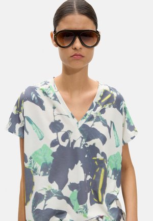 Młoda kobieta z ciemnymi włosami związanymi w kok, nosząca duże brązowe okulary przeciwsłoneczne oraz białą koszulkę z dekoltem w serek, ozdobioną zielonymi i szarymi liściastymi wzorami.