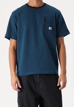 Turkusowa koszulka z krótkim rękawem wykonana z bawełny, z kieszenią na zamek błyskawiczny i czarnym ściągaczem przy szyi. Noszona z czarnymi spodniami.