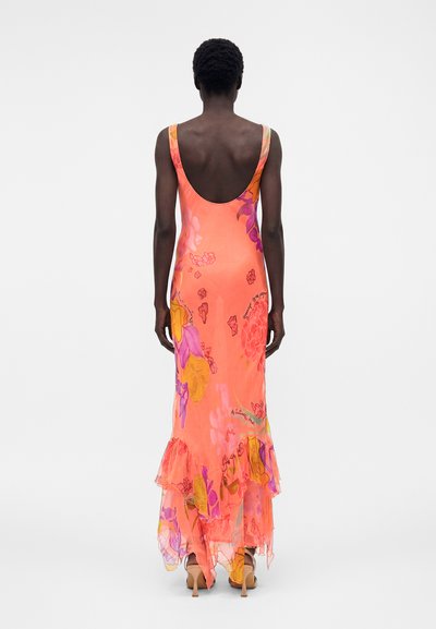 Γυναίκα όρθια με την πλάτη, φορώντας ένα πορτοκαλί μακρύ φόρεμα χωρίς μανίκια και με χαμηλή πλάτη, με φλοράλ σχέδια, βολάν στο τελείωμα και μπεζ πέδιλα με ψηλό τακούνι.