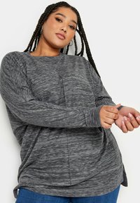 Grå långärmad topp med en texturerad, något marmorerad mönster. Har en avslappnad passform och rundad nederkant, i kombination med blå jeans.