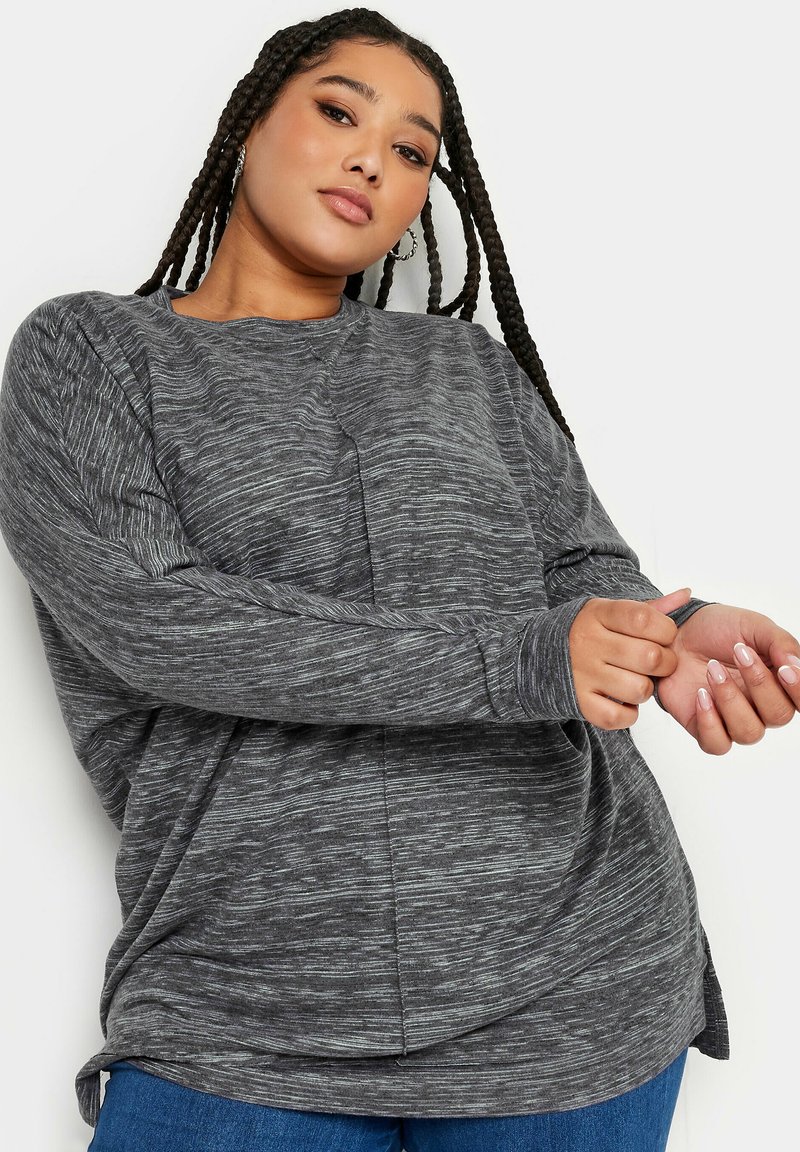 Grå långärmad topp med en texturerad, något marmorerad mönster. Har en avslappnad passform och rundad nederkant, i kombination med blå jeans.
