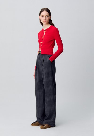 Cardigan rosso a coste con fiocchi dorati sul davanti, abbinato a pantaloni a gamba larga a righe scure a vita alta e scarpe stringate marroni.