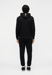 Juodas džemperis su kapišonu ir laisvalaikio kelnės. Džempyje yra raišteliai, ribinės rankogaliai ir dryžuotas akcentas nugaroje. Pagaminta iš lygaus medvilnės mišinio.