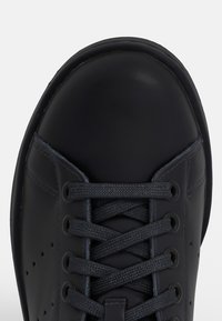 Černá kožená teniska s kulatou špičkou, texturovaným povrchem, plochými tkaničkami a perforovanými detaily po stranách.