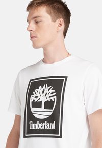 Balta medvilnės marškinėliai su dideliu juodu Timberland logotipu ir medžiu bei prekių ženklu priekyje. Trumpomis rankovėmis ir apvaliu iškirpimu.