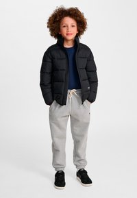 Barn med krøllet hår iført en sort vatteret jakke, marineblå skjorte, grå joggingbukser og sorte sneakers, stående med hænderne i lommerne.