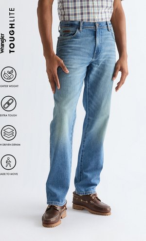 Man draagt lichtblauwe spijkerbroek, geruit overhemd in de broek gestopt en bruine leren schoenen, staand tegen een eenvoudige lichte achtergrond.