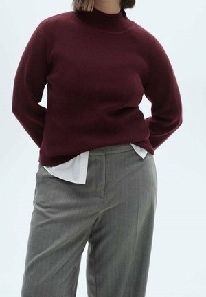 Personne portant un pull en maille bordeaux sur une chemise blanche, associé à un pantalon gris taille haute, debout les mains dans le dos.