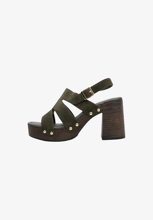 Sandalia de plataforma de ante verde oliva con tiras anchas, tachuelas doradas, hebilla ajustable en el tobillo y tacón grueso de bloque de madera marrón oscuro.