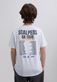 T-shirt en coton blanc avec le texte noir "SCALPERS ON TOUR" et une liste de dates de tournée au dos, avec un accent orange et un logo de crâne.