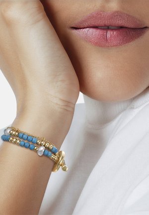 Hand van een vrouw die op haar gezicht rust, met blauwe en gouden armbanden met parelaccenten, en zachte roze lippenstift op de lippen.