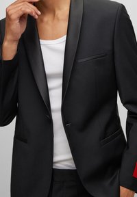 Blazer nero con revers in satin, chiusura a un bottone e due tasche frontali. Indossato sopra un top bianco, caratterizzato da un'accente rossa sul polsino.