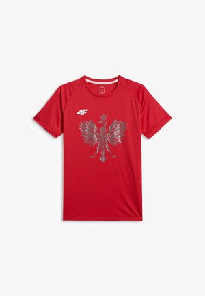Młody mężczyzna z krótkimi włosami, ubrany w czerwoną sportową koszulkę z srebrnym geometrycznym orłem i czarne spodnie na jednolitym szarym tle.