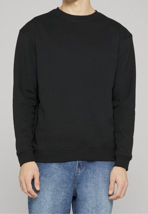 Person trägt einen einfachen schwarzen Crewneck-Sweatshirt und blaue Jeans, steht vor einem neutral grauen Hintergrund.