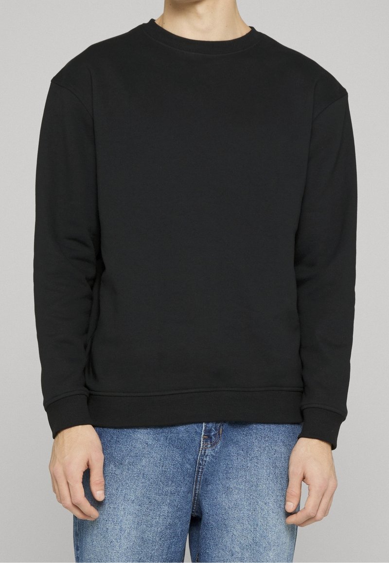 Personne portant un sweat-shirt noir uni à col rond et un jean bleu, debout devant un fond gris neutre.