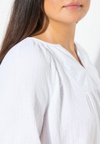Blusa bianca in lino con superficie testurizzata, scollatura a V e vestibilità ampia. Presenta dettagli arricciati sulle spalle e maniche dalla vestibilità rilassata.