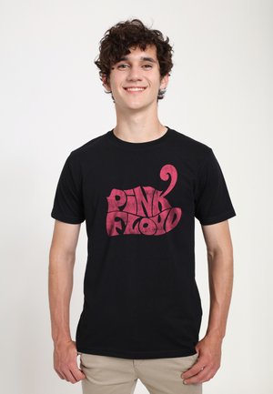 Jeune homme aux cheveux bouclés foncés, portant un t-shirt noir avec le texte rose « Pink Floyd » et un pantalon clair, debout et souriant.