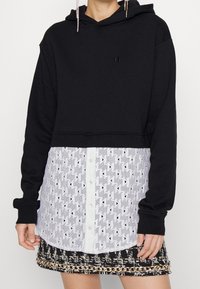 Sudadera negra cropped con una parte superior lisa y una capa inferior blanca con un diseño estampado que presenta cierre de botones y un diseño de encaje geométrico.