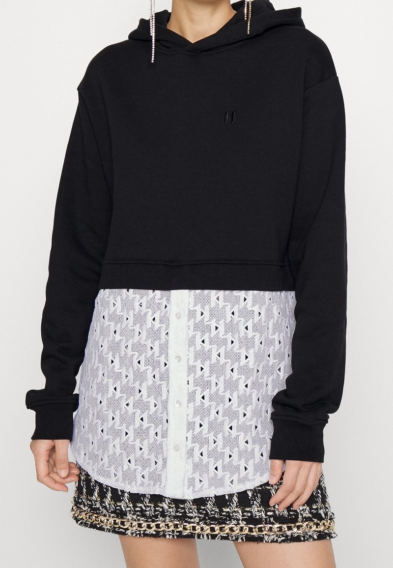 Sudadera negra cropped con una parte superior lisa y una capa inferior blanca con un diseño estampado que presenta cierre de botones y un diseño de encaje geométrico.