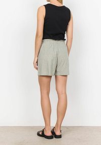 Kobieta w czarnej bezrękawnikowej bluzce, jasnoszarych, wysokich szortach i czarnych sandałach, stojąca przodem do białej ściany.