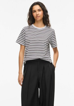 Kvinde iført en hvid og sort vandretstribet t-shirt, der er stukket ned i et par sorte højtaljede, vidde bukser med plisseringer.