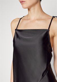 Μαύρη μεταξωτή καμισόλα με λεπτές τιράντες, διαθέτοντας ομαλή υφή και μινιμαλιστικό σχέδιο. Περιλαμβάνει ετικέτα στο πλάι που είναι ορατή στο τελείωμα.
