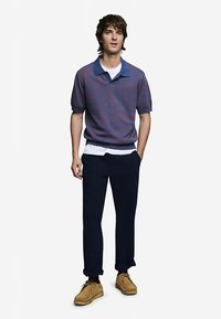 Férfi rövid ujjú, lila színű póló kék gallérral, sötét tengerészkék nadrággal és bézs cipővel párosítva. Egyszerű kialakítás, sima textúra és laza szabás.