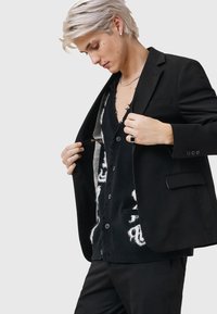 Svart blazer med en strukturerad design, bärs över en svart, mönstrad, knappad cardigan. Cardiganen har vita accenter och en djup halsringning.