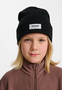 Bonnet tricoté noir avec un revers comportant une étiquette blanche portant l'inscription "hummel sportswear". Porté sur une veste zippée en polaire marron.