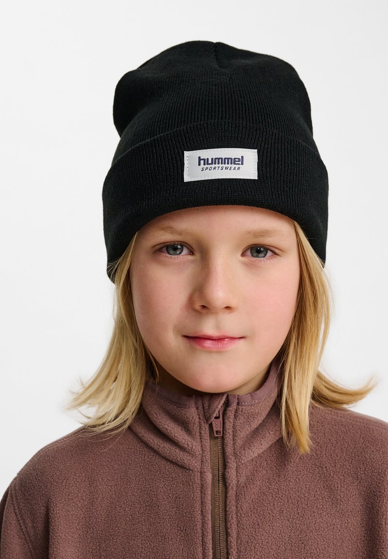 Bonnet tricoté noir avec un revers comportant une étiquette blanche portant l'inscription "hummel sportswear". Porté sur une veste zippée en polaire marron.