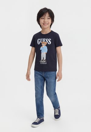 Ragazzo giovane che cammina, indossando una t-shirt blu navy con la grafica di un orsetto e la scritta "GUESS", jeans blu e scarpe da ginnastica blu navy con lacci bianchi.