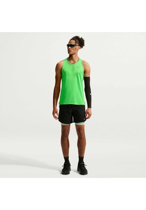Atletische man draagt een groen mouwloos shirt, zwarte korte broek met groene bies, zwarte compressie-armmouw, zwarte sokken, schoenen en zonnebril staand.